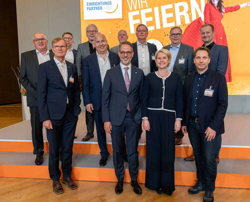 Gruppe von Geschäftsleuten in Anzügen und Businesskleidung posiert lächelnd auf einer Bühne vor einer orangefarbenen Jubiläumswand mit dem Schriftzug „Einrichtungspartner Ring – Wir feiern“