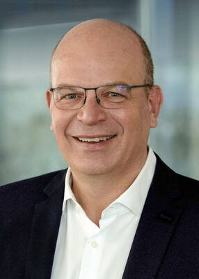 Lächelnder, glatzköpfiger Mann mit Brille im dunklen Sakko und weißem Hemd vor unscharfem Hintergrund