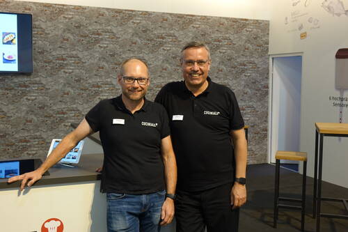 Zwei lächelnde Mitarbeiter im schwarzen Cuciniale-Poloshirt stehen nebeneinander an einem Messestand mit Backsteinwand, Tablets und Stehtischen im Hintergrund