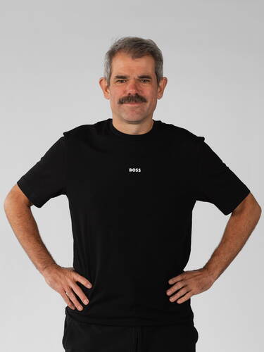 Mittelalterlicher Mann mit Schnurrbart steht selbstbewusst mit in die Hüften gestemmten Händen in schwarzem T-Shirt vor neutralem Hintergrund