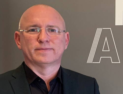 Glatzköpfiger Mann mit Brille im dunklen Anzug vor grauem Hintergrund mit großem Buchstaben „A“