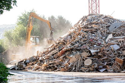 Bagger mit Greifarm sortiert einen riesigen Haufen aus Holz- und Bauschutt unter staubiger Luft auf einem Recyclinghof