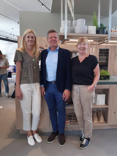 Drei lächelnde Personen stehen nebeneinander in einer modernen Küche mit Holzregalen und dekorativen Gefäßen im Hintergrund.