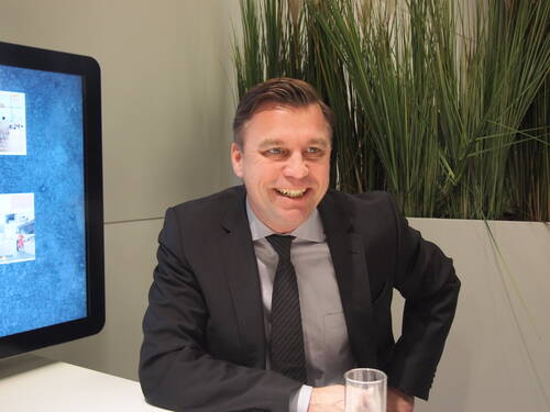 Lächelnder Mann im Anzug sitzt an einem weißen Tisch in modernem Büroambiente neben einem Bildschirm und hohen Ziergräsern