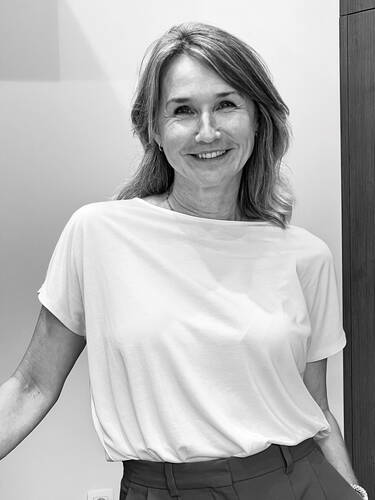 Lächelnde Frau mittleren Alters in schlichtem weißen T‑Shirt lehnt entspannt vor einer hellen Wand und blickt direkt in die Kamera