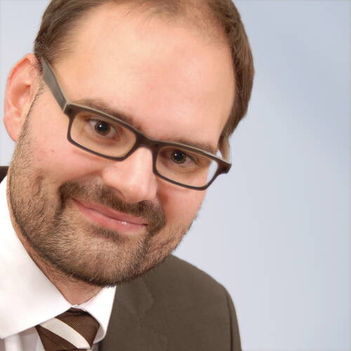 Mann mit Brille, Bart und Anzug lächelt schräg in die Kamera vor hellem Hintergrund