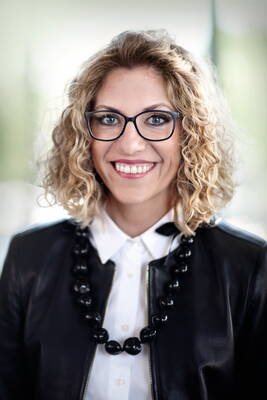 Lächelnde Frau mit lockigen blonden Haaren, schwarzer Brille, weißer Bluse und schwarzer Lederjacke vor unscharfem, hellgrünem Hintergrund
