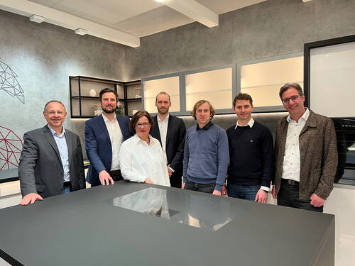 Sieben Personen stehen lächelnd nebeneinander hinter einer modernen Kücheninsel in einem hell beleuchteten Showroom mit grauer Wand und offenen Regalen.