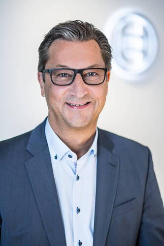 Geschäftsmann mittleren Alters mit Brille und grauem Anzug lächelt vor hellem Hintergrund mit unscharfem Firmenlogo.