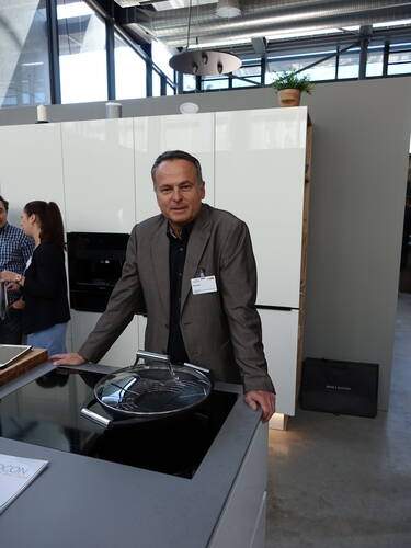 Mann im Anzug lehnt sich in moderner Ausstellungsküche über ein Kochfeld mit Glasdeckel, während im Hintergrund weitere Personen in dem lichtdurchfluteten Raum sprechen