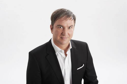 Mittelalter Mann in schwarzem Anzug und weißem Hemd steht vor hellem Hintergrund und blickt lächelnd in die Kamera