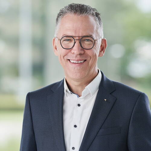 Lächelnder Mann mittleren Alters mit Brille und Anzugjacke steht vor unscharfem, hellem Hintergrund