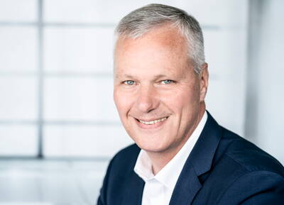 Älterer Mann mit grauen Haaren im dunklen Anzug lächelt freundlich in die Kamera vor einem hellen, unscharfen Bürohintergrund.