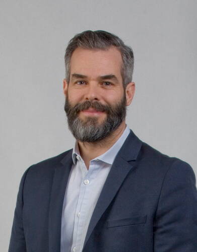 Mittelalterlicher Mann mit graumelierter Kurzhaarfrisur und Vollbart lächelt dezent in die Kamera und trägt ein dunkelblaues Sakko über einem hellen Hemd vor neutralem Hintergrund.