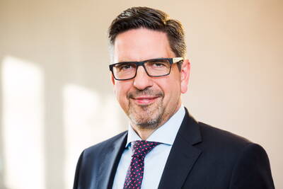 Mittelalterlicher Mann mit Brille und Bart lächelt im Anzug und gemusterter Krawatte vor einem hellen, unscharfen Hintergrund