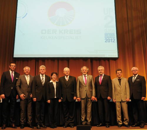 Gruppe von neun elegant gekleideten Personen steht lächelnd auf einer Bühne vor einer großen Leinwand mit dem Logo „DER KREIS Keukenspecialist“ und der Aufschrift „UP 2 date DER KREIS KONGRESS 2012“