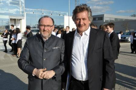 Zwei mittelalte Männer in Anzügen stehen im Sonnenschein vor einer modernen Glasfassade und lächeln in die Kamera, während im Hintergrund mehrere Personen in Businesskleidung miteinander sprechen.