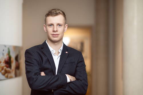 Junger Mann im dunkelblauen Anzug mit verschränkten Armen steht selbstbewusst in einem hellen Büroflur