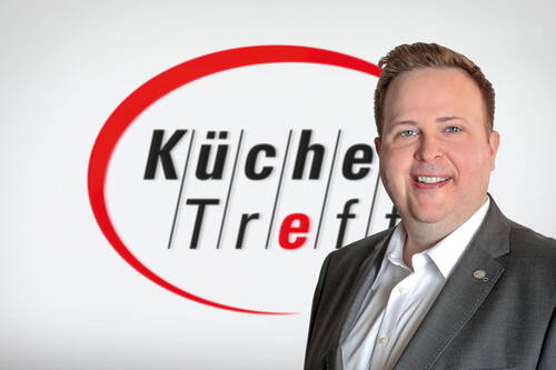 Lächelnder Mann im Anzug steht vor einem weißen Hintergrund mit dem rot-schwarzen Logo „Küchen Treff“ im Halbkreis-Design