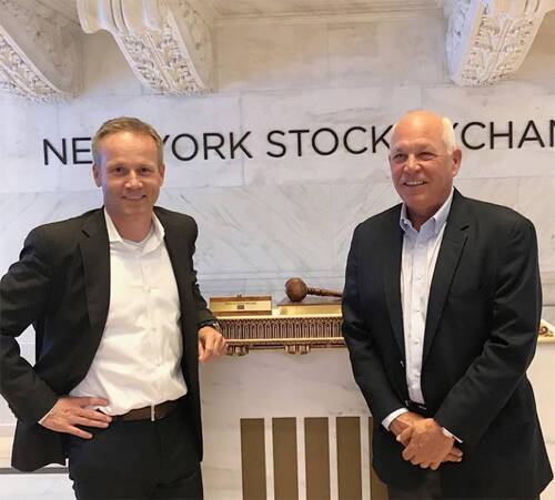 Zwei Männer im Anzug stehen lächelnd in der Eingangshalle der New York Stock Exchange vor einem Geländer mit Richterhammer.