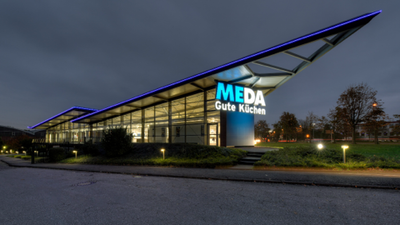 Modernes Küchenstudio von „MEDA Gute Küchen“ mit spitz zulaufendem Glasdach und blauer LED-Beleuchtung in der Abenddämmerung