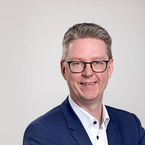 Lächelnder Mann mit Brille in dunkelblauem Anzug und weißem Hemd vor neutral hellem Hintergrund