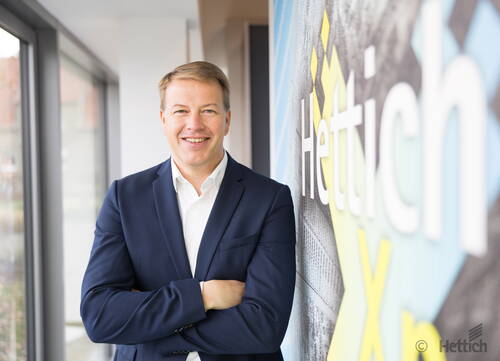 Lächelnder Geschäftsmann im dunkelblauen Anzug steht mit verschränkten Armen in einem modernen Büroflur neben einer Wand mit großem Hettich-Logo.