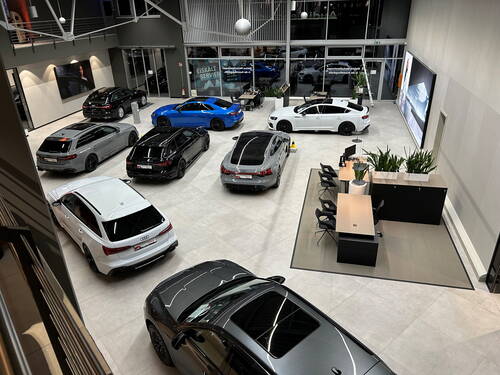 Moderne Audi-Ausstellungshalle mit mehreren sportlichen Limousinen und Kombis in verschiedenen Farben, geparkten Fahrzeugreihen und einem Beratungsbereich mit Schreibtischen und Pflanzen im Vordergrund