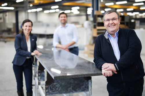 Drei lächelnde Personen in Businesskleidung stehen in einer Werkhalle an einer polierten Steinplatte und präsentieren gemeinsam das fertige Werkstück