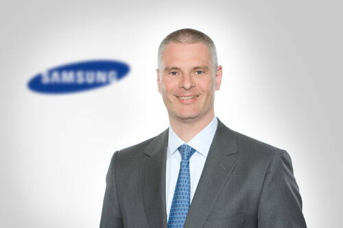 Lächelnder Geschäftsmann im grauen Anzug und blauer Krawatte vor einem unscharfen Samsung-Logo auf hellem Hintergrund