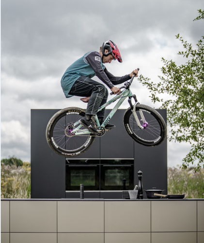 Mountainbiker springt mit seinem Fahrrad spektakulär über eine moderne Outdoor-Küche vor wolkenverhangenem Himmel.