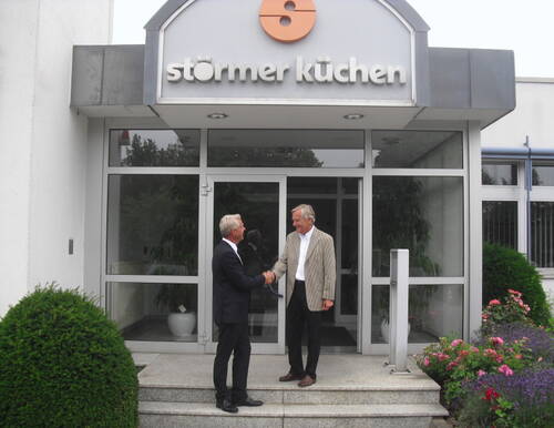Zwei Männer im Anzug begrüßen sich per Handschlag vor dem Eingangsbereich der Firma „Störmer Küchen“