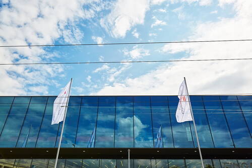 Moderne Glasfassade mit spiegelndem Himmel und zwei wehenden weißen Fahnen vor dem Gebäude