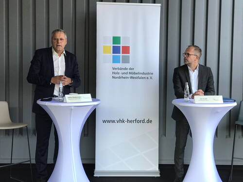 Zwei Herren im Anzug stehen an hohen Stehtischen neben einem Roll-up der Verbände der Holz- und Möbelindustrie Nordrhein-Westfalen und sprechen bei einer Konferenz oder Presseveranstaltung.