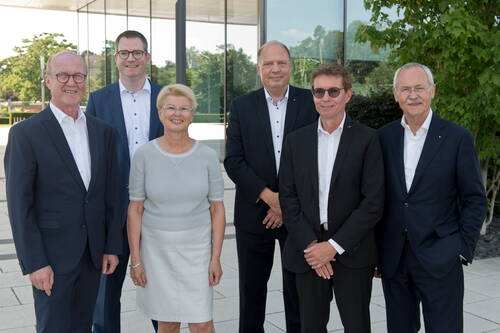 Sechs ältere Geschäftspersonen in formeller Kleidung stehen lächelnd nebeneinander vor einem modernen Bürogebäude mit Glasfassade im Grünen.