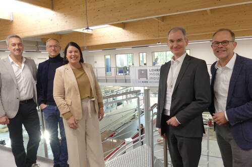 Fünf lächelnde Geschäftsleute stehen in moderner Ausbildungswerkstatt mit Holzbalkendecke und Geländer, im Hintergrund ein Banner mit der Aufschrift „Lehrfabrik Möbelindustrie“.