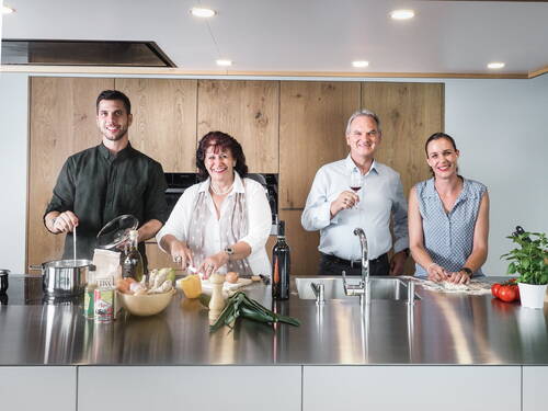 Vier Erwachsene kochen und backen gemeinsam an einer modernen Kücheninsel, umgeben von frischem Gemüse, Kräutern, Öl und Wein, während sie lächeln und miteinander interagieren.