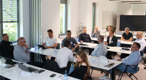 Teilnehmende sitzen in einem modernen Konferenzraum an U-förmig angeordneten Tischen, arbeiten an Laptops und diskutieren konzentriert während eines geschäftlichen Workshops.