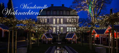 Festlich beleuchtetes Herrenhaus mit Lichterketten, kleinen Weihnachtsbuden und der Aufschrift „Wundervolle Weihnachten!“ in der Abenddämmerung