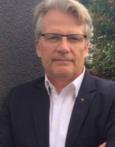Mittelälterlicher Mann mit grauem Haar und Brille steht im Anzug vor einer dunklen Wand und blickt ernst in die Kamera