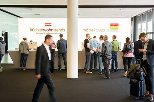 Besucher stehen an einem langen Empfangstresen der Fachmessen „küchenwohntrends“ und „möbel austria“, während andere Personen mit Koffern und Smartphones durch die moderne Messehalle gehen.