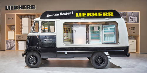 Historischer Liebherr-Lieferwagen mit seitlich eingebauten Kühlschränken steht in einer Messeausstellung vor einer Wand mit Kühlschrankmodellen aus verschiedenen Jahrzehnten.