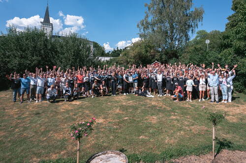 Große Menschengruppe feiert im Freien auf einer Wiese, hebt jubelnd die Arme und präsentiert gemeinsam eine Auszeichnung vor grünen Bäumen und blauem Himmel.