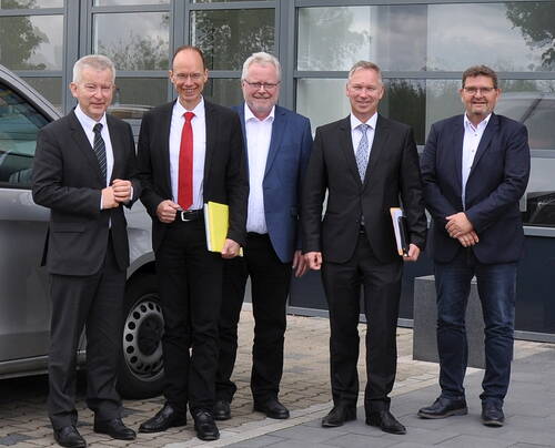 Fünf Männer in Anzügen stehen lächelnd nebeneinander vor einem Firmengebäude und halten Unterlagen in der Hand
