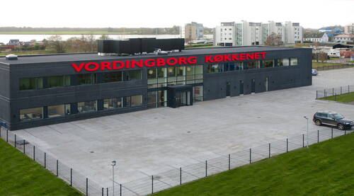 Modernes Firmengebäude von „Vordingborg Køkkenet“ mit großer Glasfassade, weiträumigem Parkplatz und umliegenden Wohnhäusern im Hintergrund