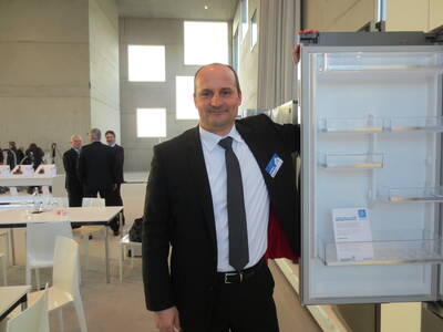 Geschäftsmann im Anzug präsentiert in einer modernen Messehalle einen geöffneten, leeren Kühlschrank mit Informationsblatt in der Tür