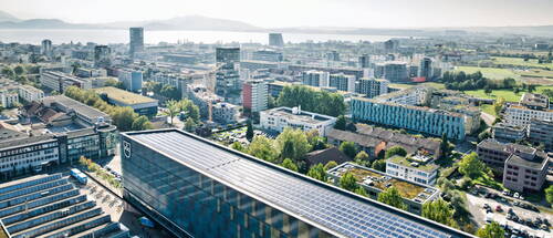 Solaranlage auf dem Dach eines modernen Stadtgebäudes inmitten dichter Wohn- und Bürohäuser mit See und Hügeln im Hintergrund