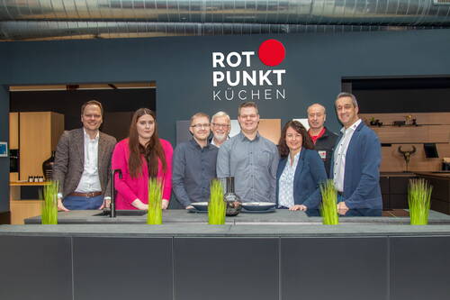 Achtköpfiges Team steht in einer modernen Ausstellungsküche vor dem Logo von Rotpunkt Küchen hinter einer breiten Kochinsel mit Dekogräsern und Geschirr