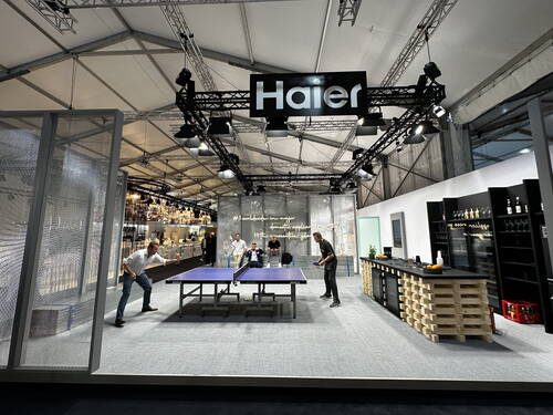 Mehrere Personen spielen Tischtennis in einem modern gestalteten Haier-Messestand mit beleuchtetem Firmenlogo, offener Küchenzeile und Barbereich im Hintergrund.