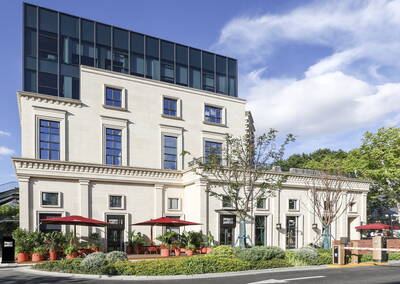 Modernes Stadthaus mit heller Natursteinfassade, großflächiger Glasetage im Obergeschoss und straßenseitiger Terrasse mit roten Sonnenschirmen und üppigen Pflanzkübeln an einem sonnigen Tag.